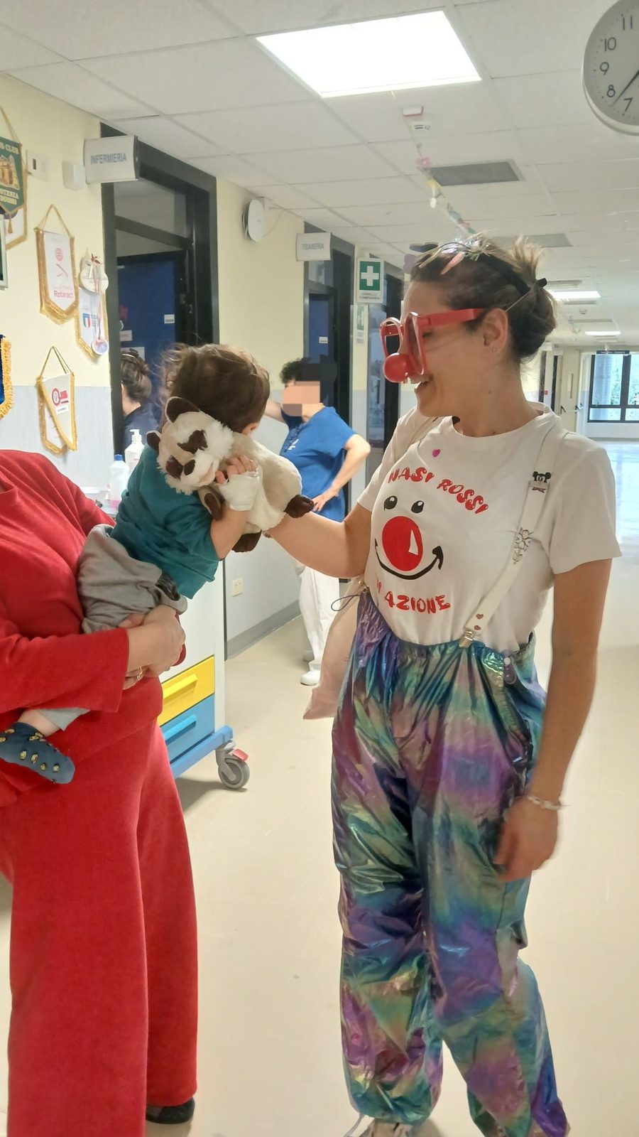 Una clown volontaria gioca con un pupazzo davanti a un bambino, nel corridoio di un reparto