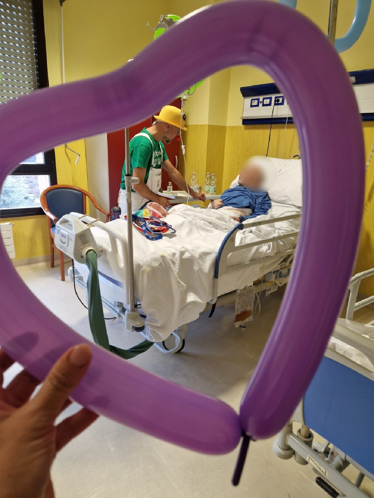 Un palloncino a forma di cuore inquadra un clown volontario accanto al letto di un piccolo paziente