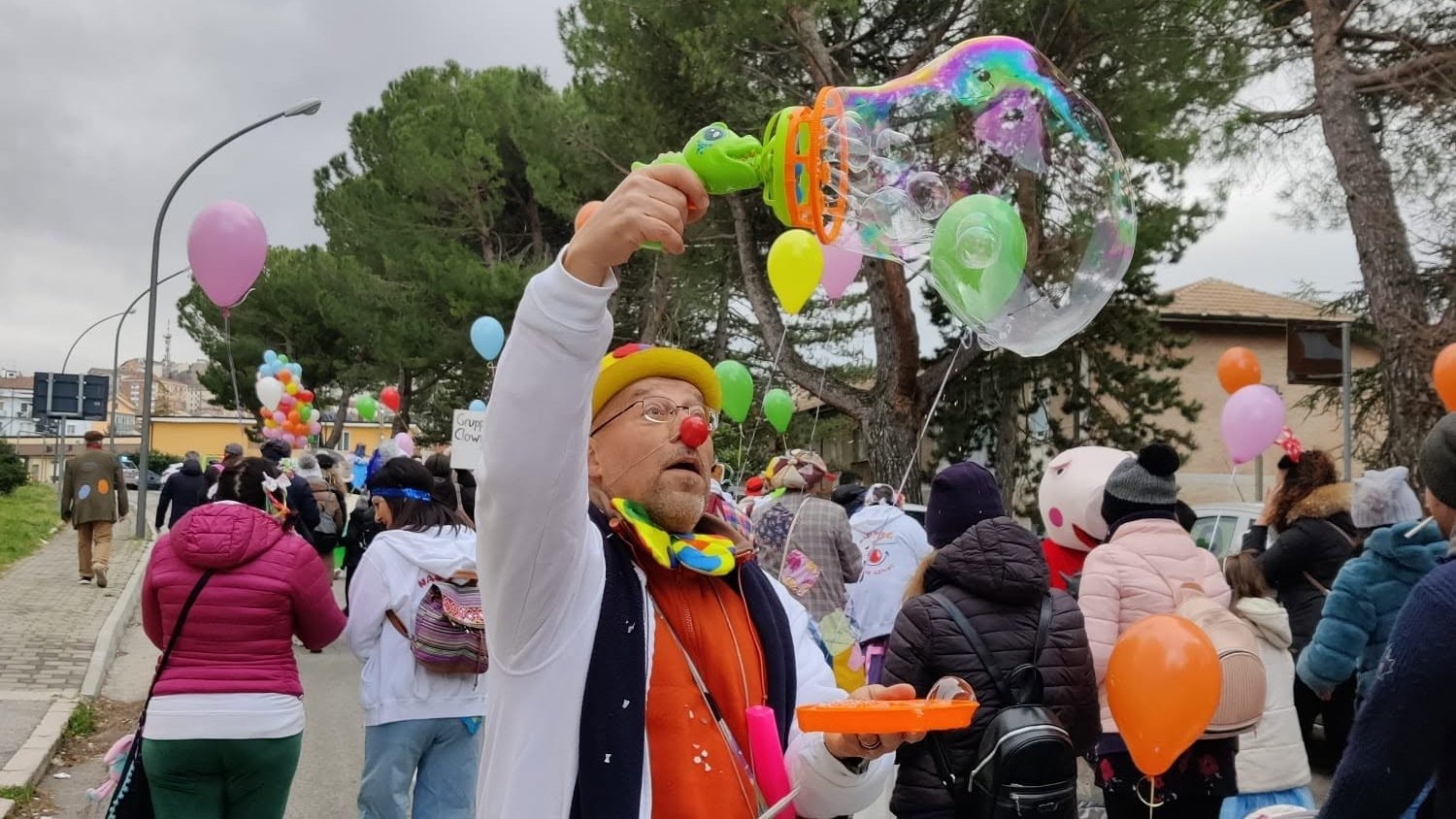 Un clown volontario fa una grande bolla di sapone durante una sfilata per le vie della città, tra palloncini e famiglie