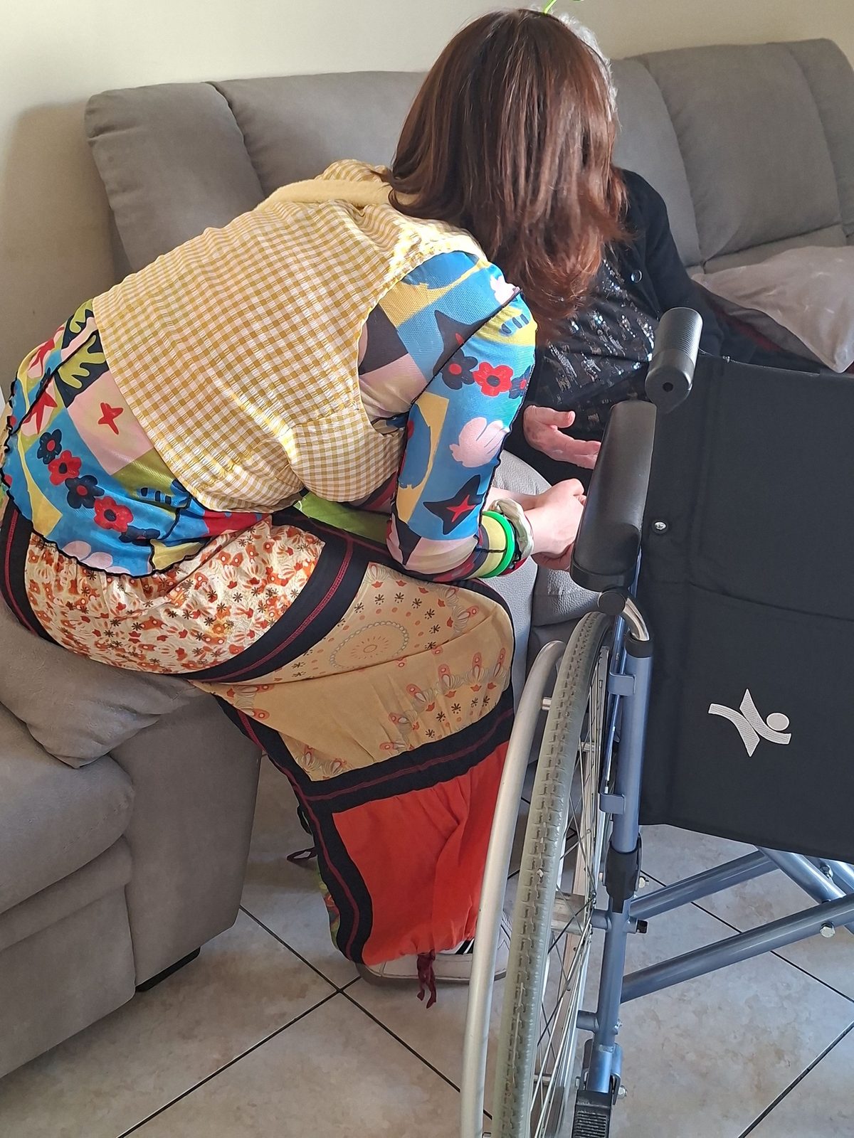 Una clown volontaria accovacciata accanto a un'anziana in carrozzina, in ascolto e mano nella mano